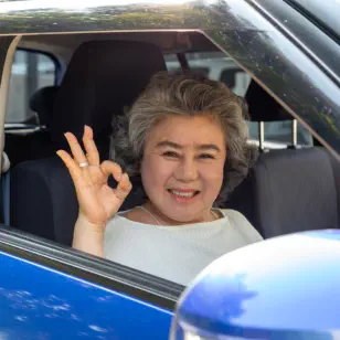 An old woman sitting inside the car smiling while doing the okay pose