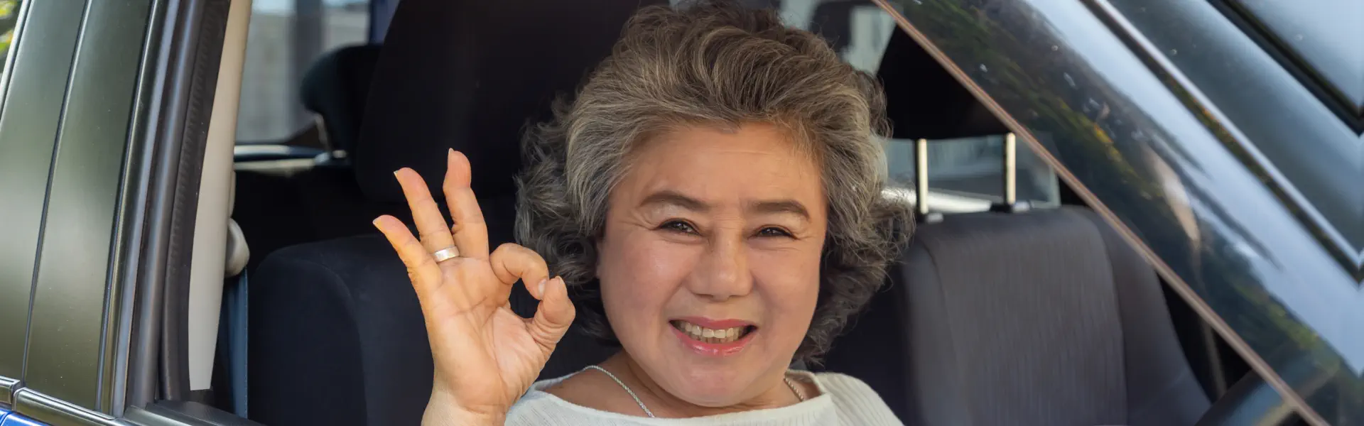 An old woman sitting inside a car and smiling while doing the okay pose